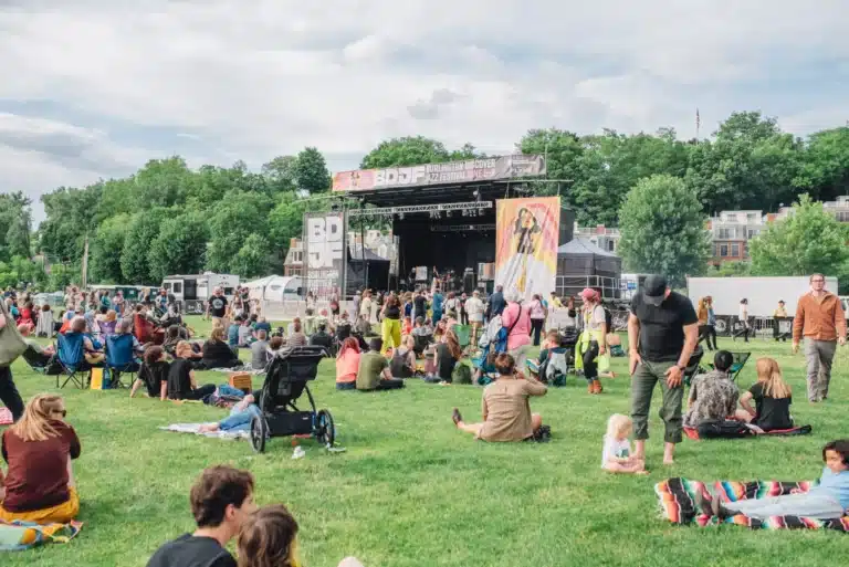 A crowd of people relax on a grassy lawn in front of an outdoor stage at the Burlington Discover Jazz Festival. Some sit on blankets or lawn chairs while others stand or walk around. The stage features banners with the festival name and artwork, and is surrounded by trees and buildings under a partly cloudy sky.