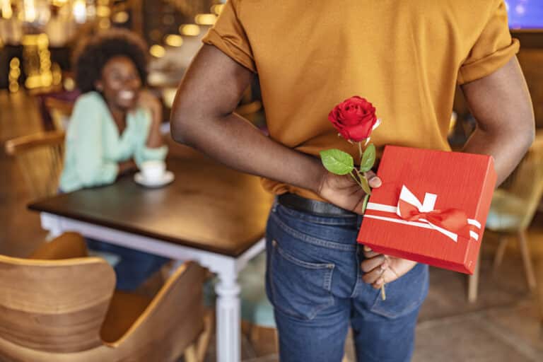 Man hiding red rose behind his back and gift for his smiling girlfriend while sitting in restaurant.